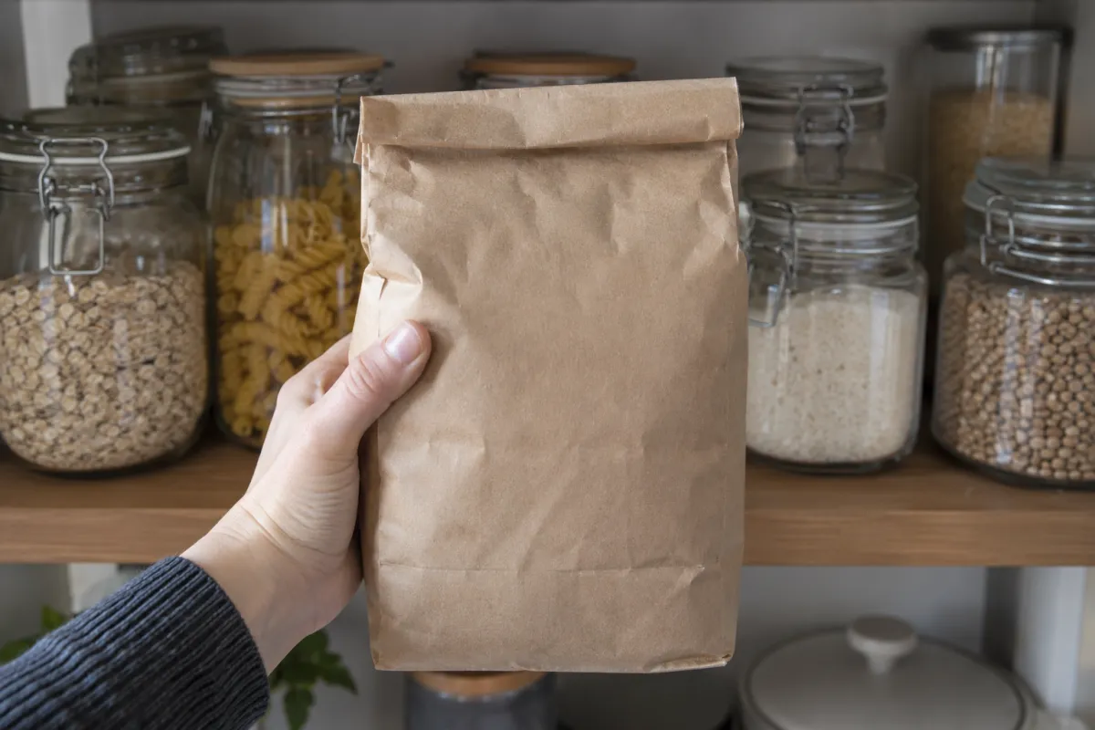 Hand holding a bag of flour in front of a pantry shelf lined with sealed glass jars
