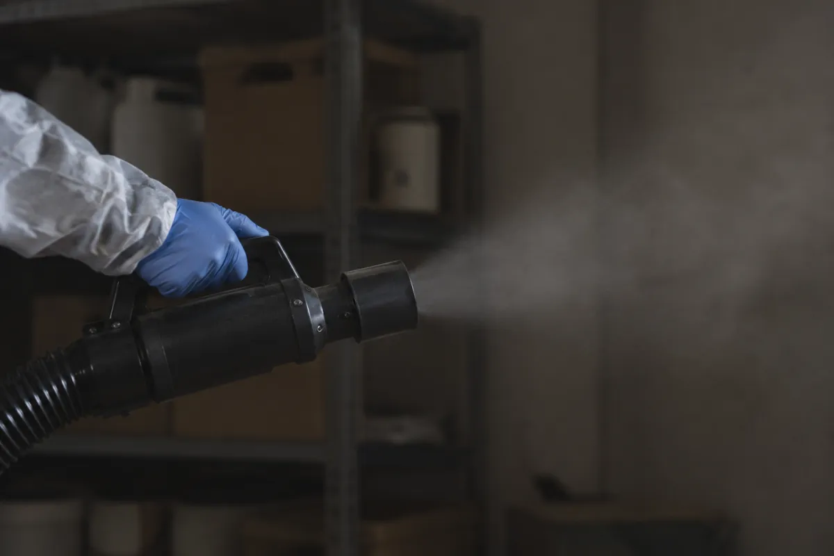 A gloved hand holding the nozzle of a fogging machine as it releases a fine mist in a dim storeroom